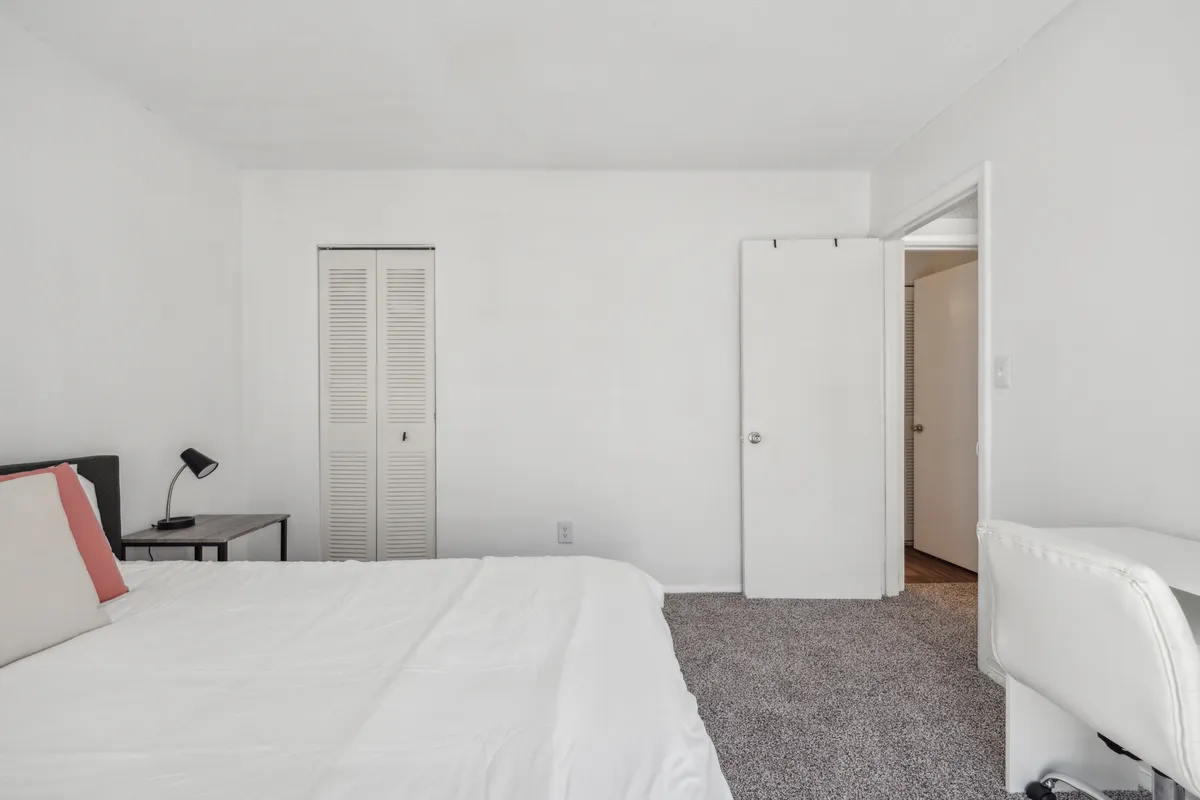 This image features a tranquil bedroom scene showcasing a comfortable bed with inviting bedding, a nightstand, and a glimpse of the closet space. The room is softly lit by natural light, highlighting a sense of serenity and relaxation. The visible closet helps communicate the promise of ample storage, while the overall layout expresses a welcoming, peaceful atmosphere.