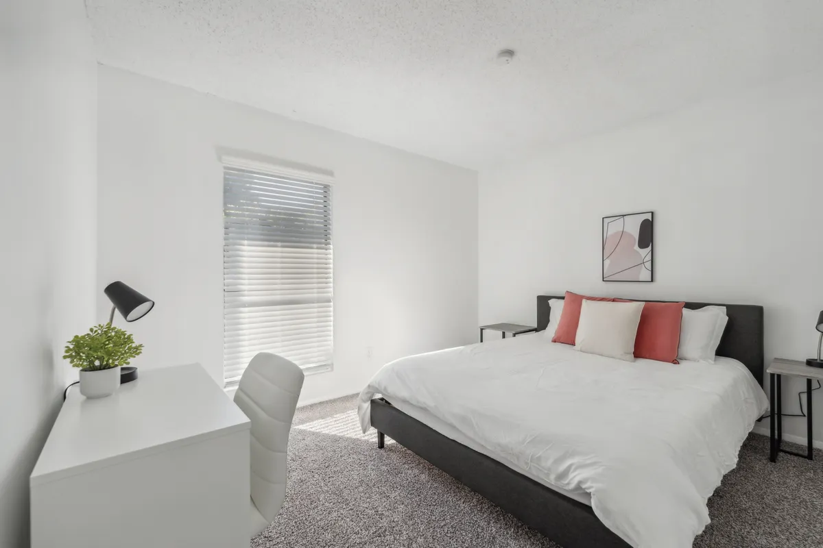 This image features a serene bedroom with white bedding, decorative pillows, a neat desk with a lamp, and a window with blinds. The calm and inviting mood suggests a peaceful retreat for guests to relax, read, or work after a day out in the city.