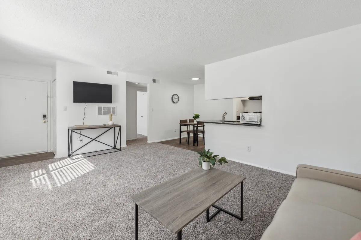 This image showcases a spacious living area with a plush sofa, coffee table, television, and a glimpse of the adjacent dining area. The space is bathed in natural light, creating an inviting atmosphere ideal for both relaxation and entertaining. The arrangement of furniture encourages socializing or solo downtime.