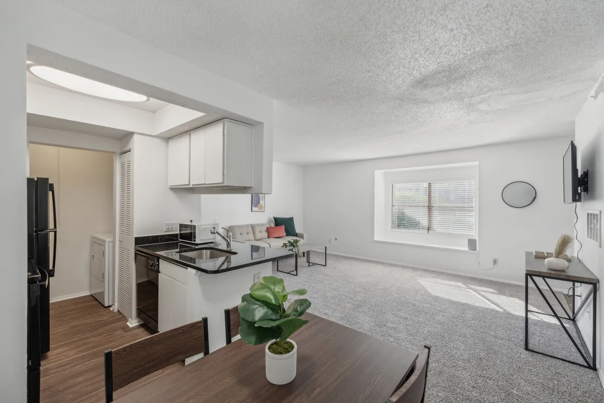 A modern kitchen and dining space with a bright kitchen island, a welcoming seating area, large windows allowing natural light, a decorative plant, and a dining table. The arrangement is perfect for sunny breakfasts or evening gatherings.